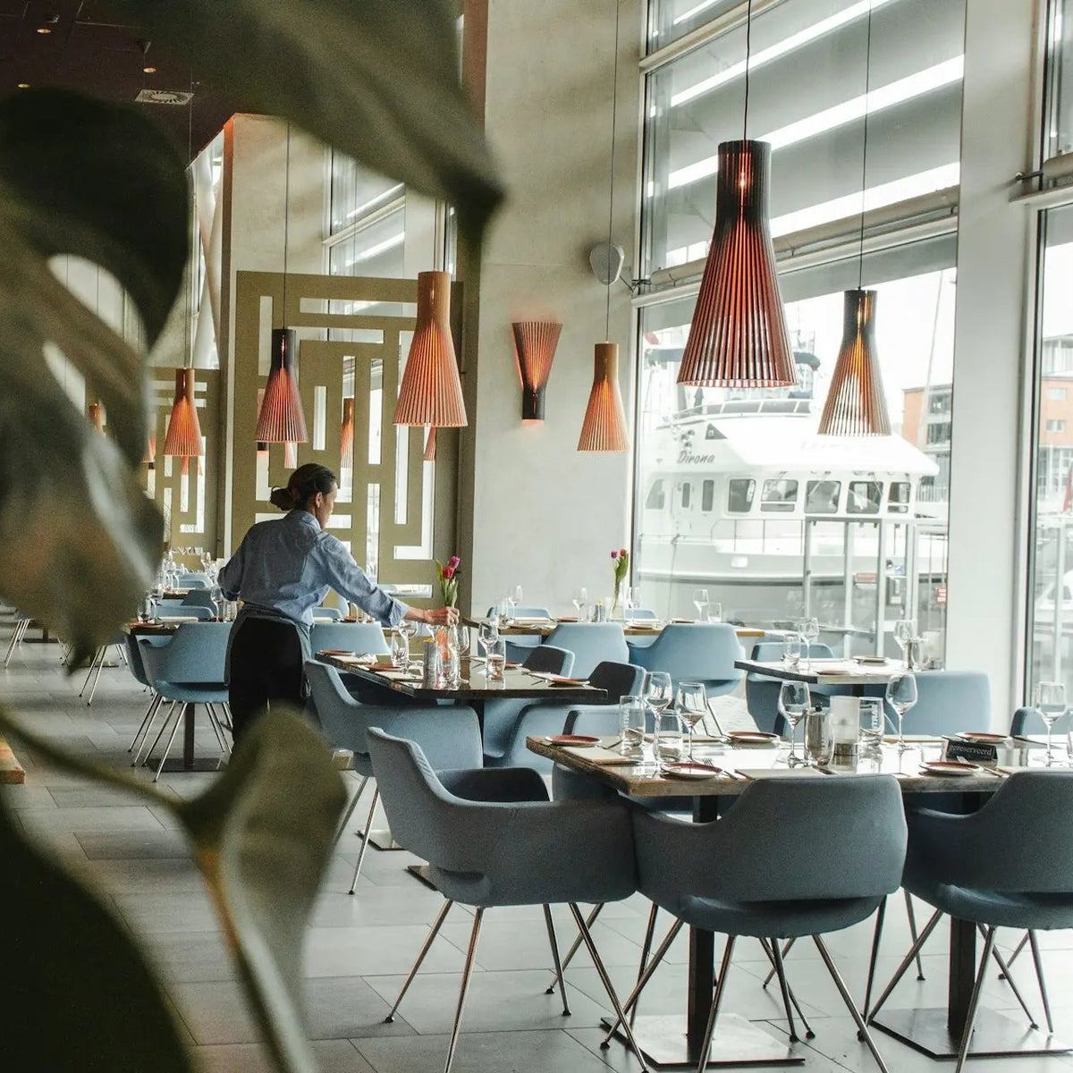 A modern restaurant interior with blue chairs, dark tables, orange pendant lights, and a person standing near the tables. Large green leaves in the foreground partially obscure the view.
