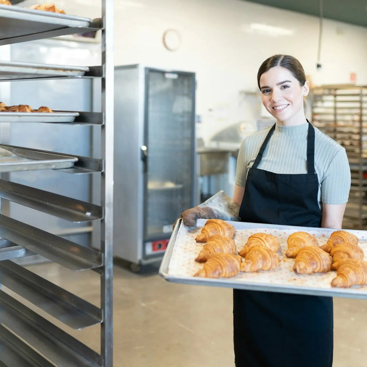 A smiling woman in a bakery wearing a black apron over a light blue shirt, holding a tray of golden-brown croissants. She is standing in a commercial bakery next to a large metal rack filled with baking trays.