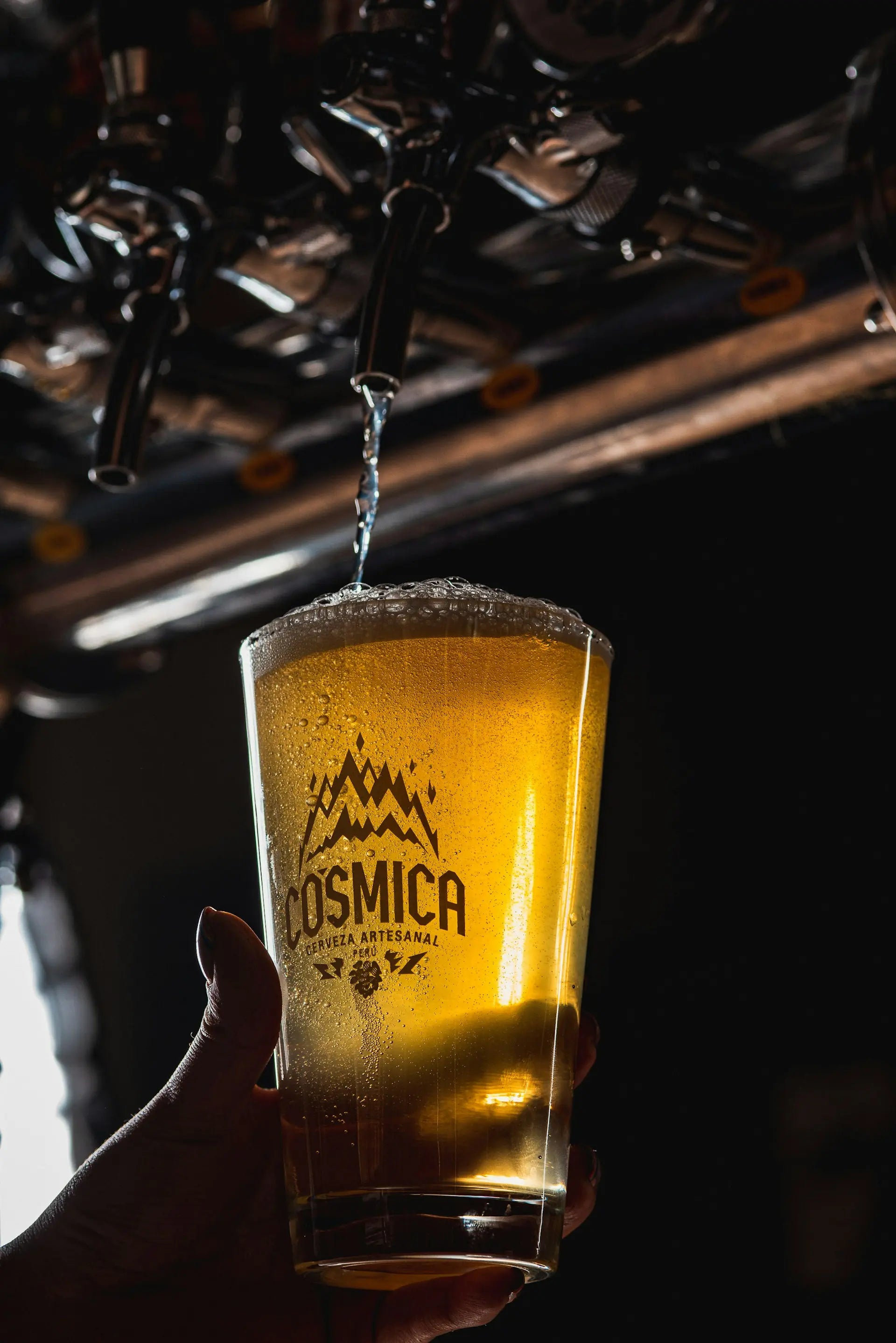A hand holds a frosted glass under a beer tap, with golden beer pouring into it. The glass is printed with the brand name 'COSMICA'.