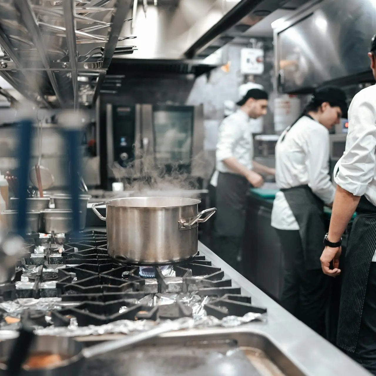 A commercial kitchen scene with several chefs in white uniforms and black aprons working around stainless steel stovetops, a large pot on a burner, and kitchen equipment in the background.