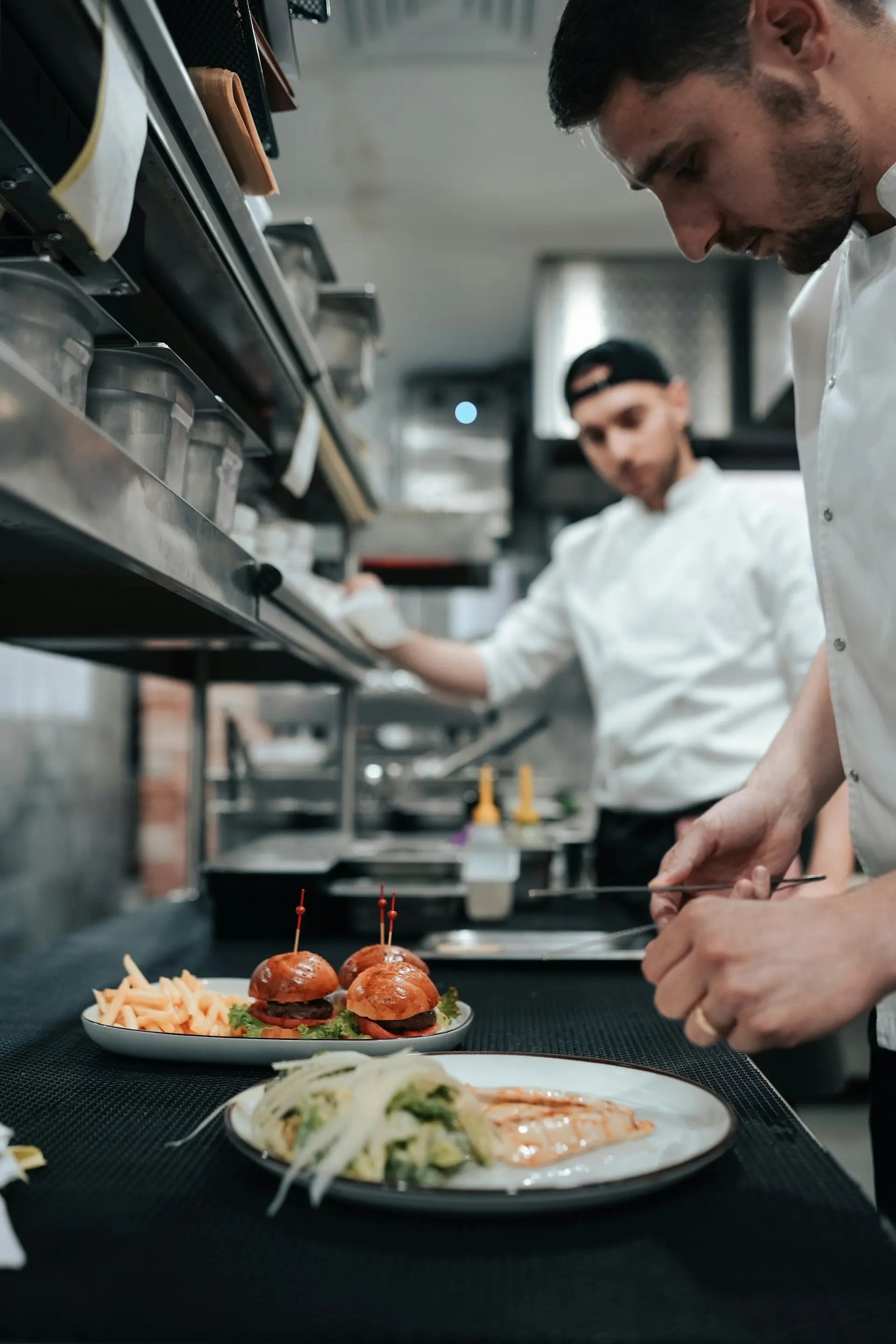 A professional kitchen scene with two chefs at work, with plated food in the foreground including sliders and a salad.