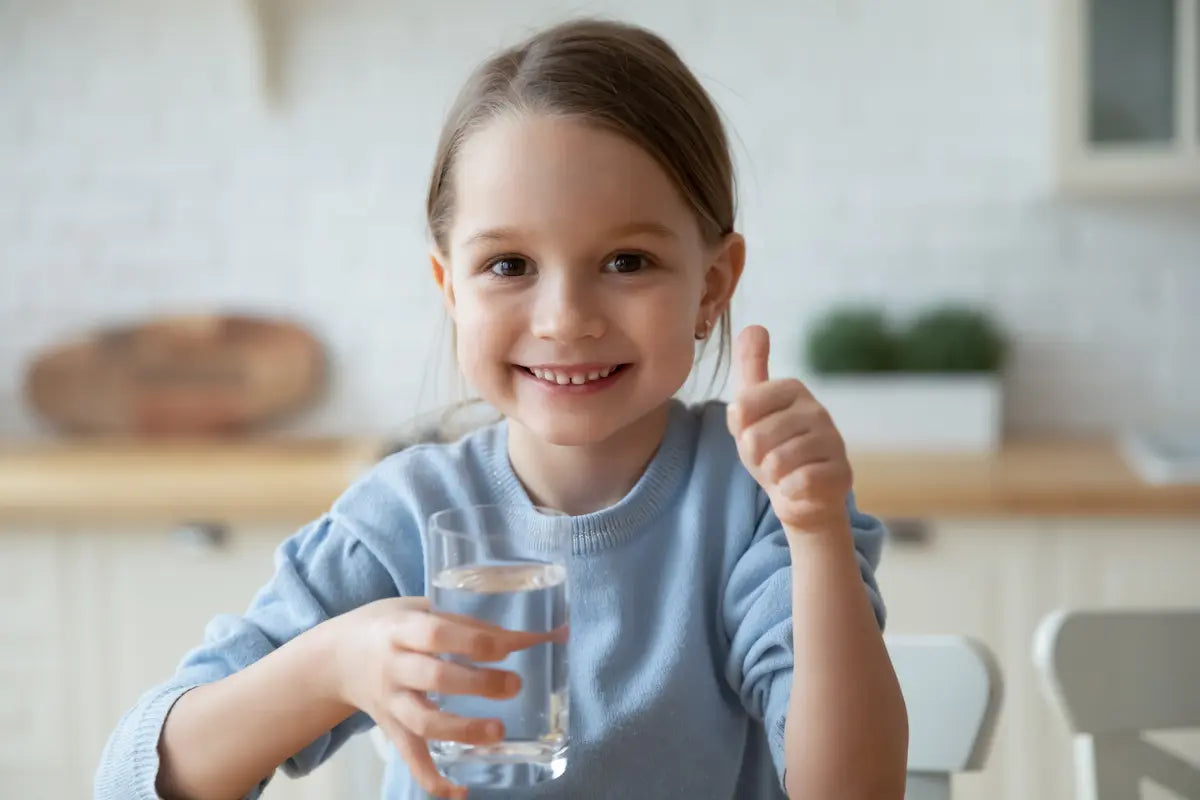 Child holding a glass of water and giving a thumbs-up in a kitchen setting